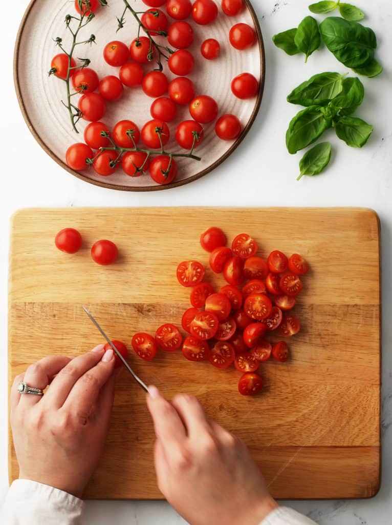 Slicing cherry tomatoes in half on a wooden cutting board with fresh basil leaves in the corner.