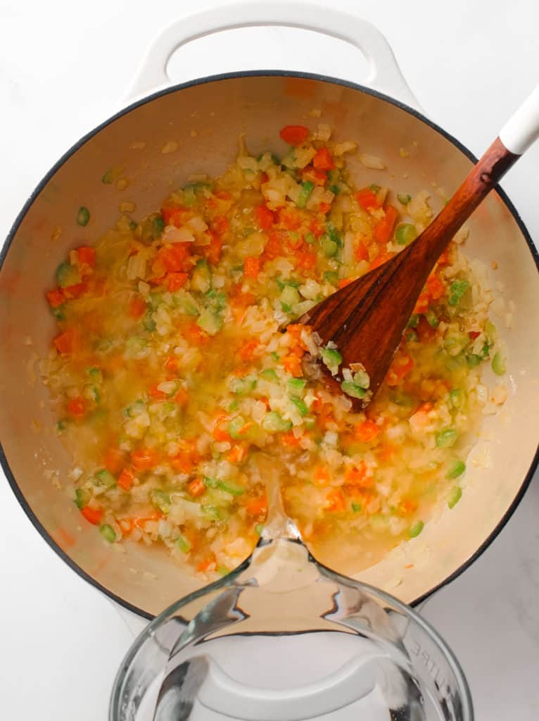 A large measuring cup pouring water into large Dutch oven pot of chopped vegetables.