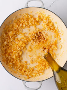 Pouring white wine in a large cooking pan with onions and garlic.