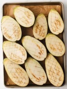 Eggplant laying on a baking tray, cut in half and sliced with a checkered pattern.