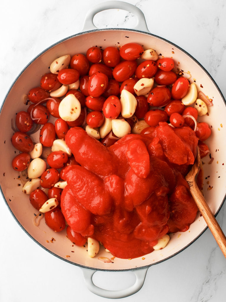 Large cooking pan with fresh tomatoes, canned tomatoes, garlic cloves and seasonings.