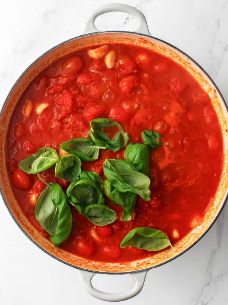 Large cooking pan with crushed tomatoes, garlic cloves and fresh basil leaves.