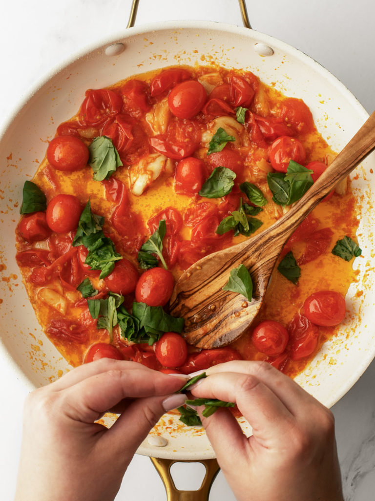 Burst tomatoes and fresh basil cooking in large pan.